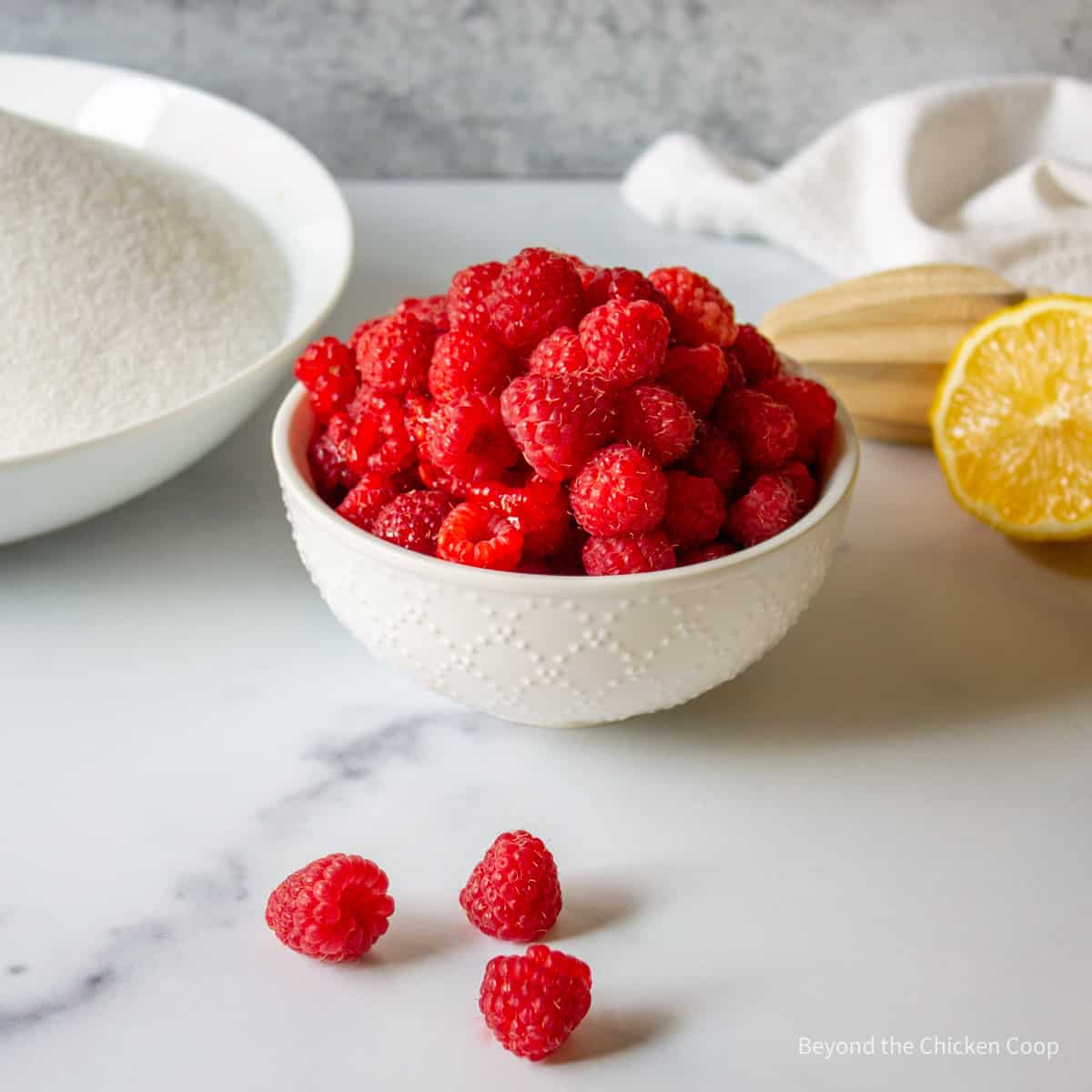 A white bowl filled with fresh raspberries next to a fresh lemon half and a bowl of sugar. 