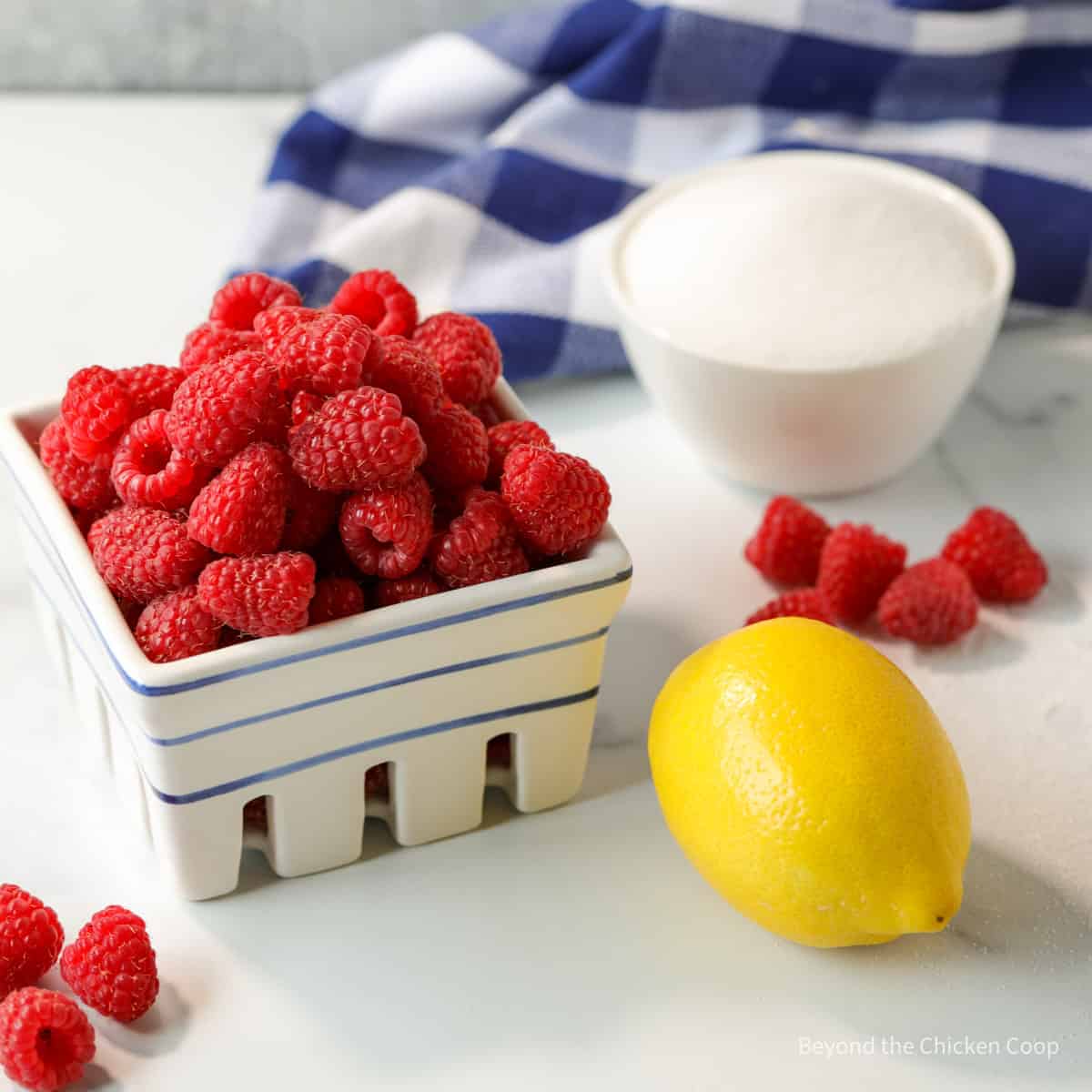 A basket with raspberries next to a fresh lemon and a bowl of sugar. 
