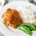 A chicken thigh with a sauce on a plate with rice and peas.