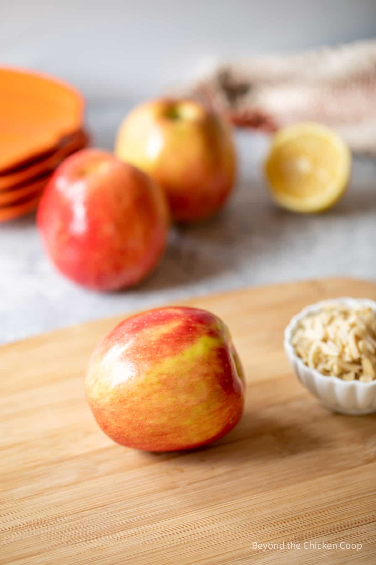 A apple on a cutting board next to a bowl of slivered almonds.