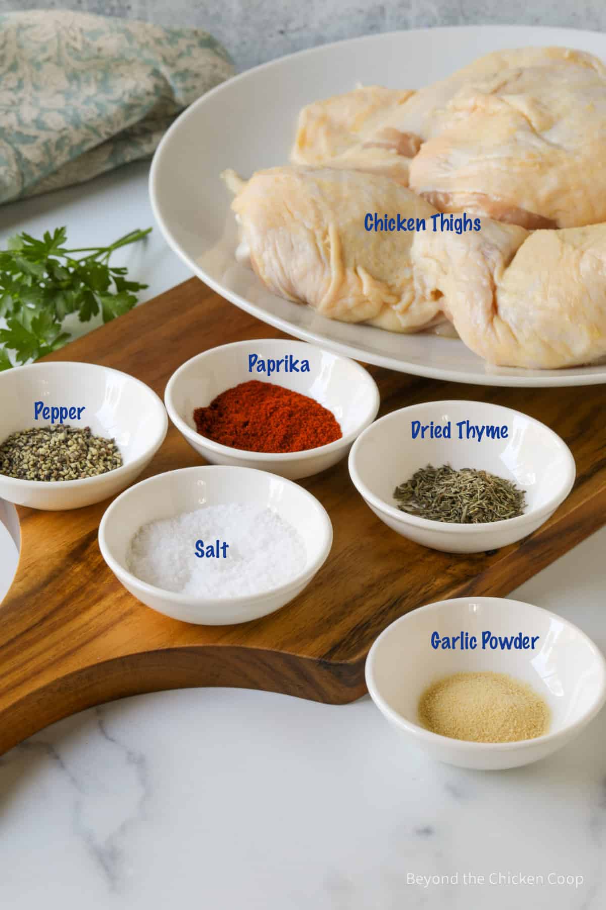 Small white bowls filled with herbs and spices next to a plate with chicken thighs. 