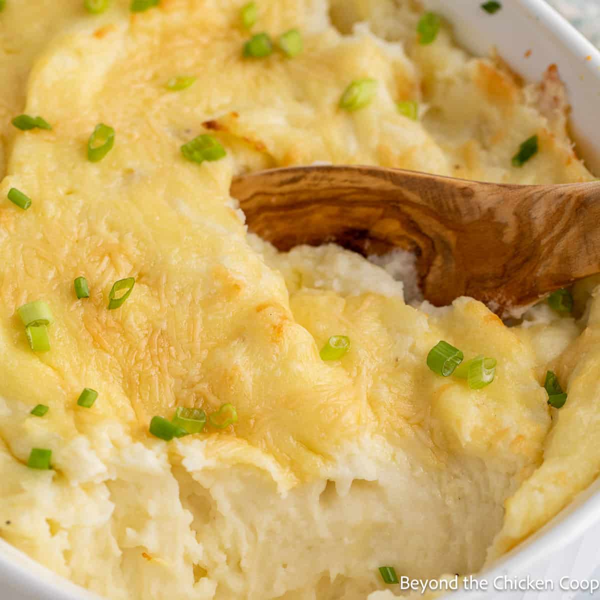 A wooden spoon scooping up mashed potatoes in a white casserole dish.