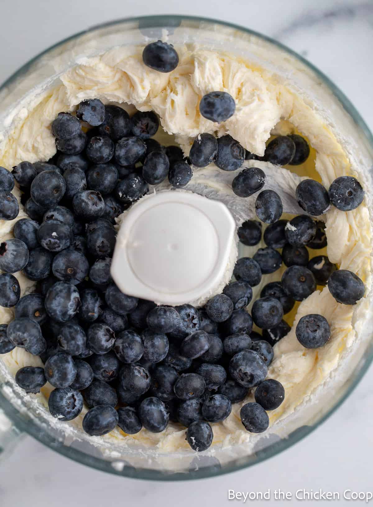 Blueberries added to a food processor filled with cream cheese.