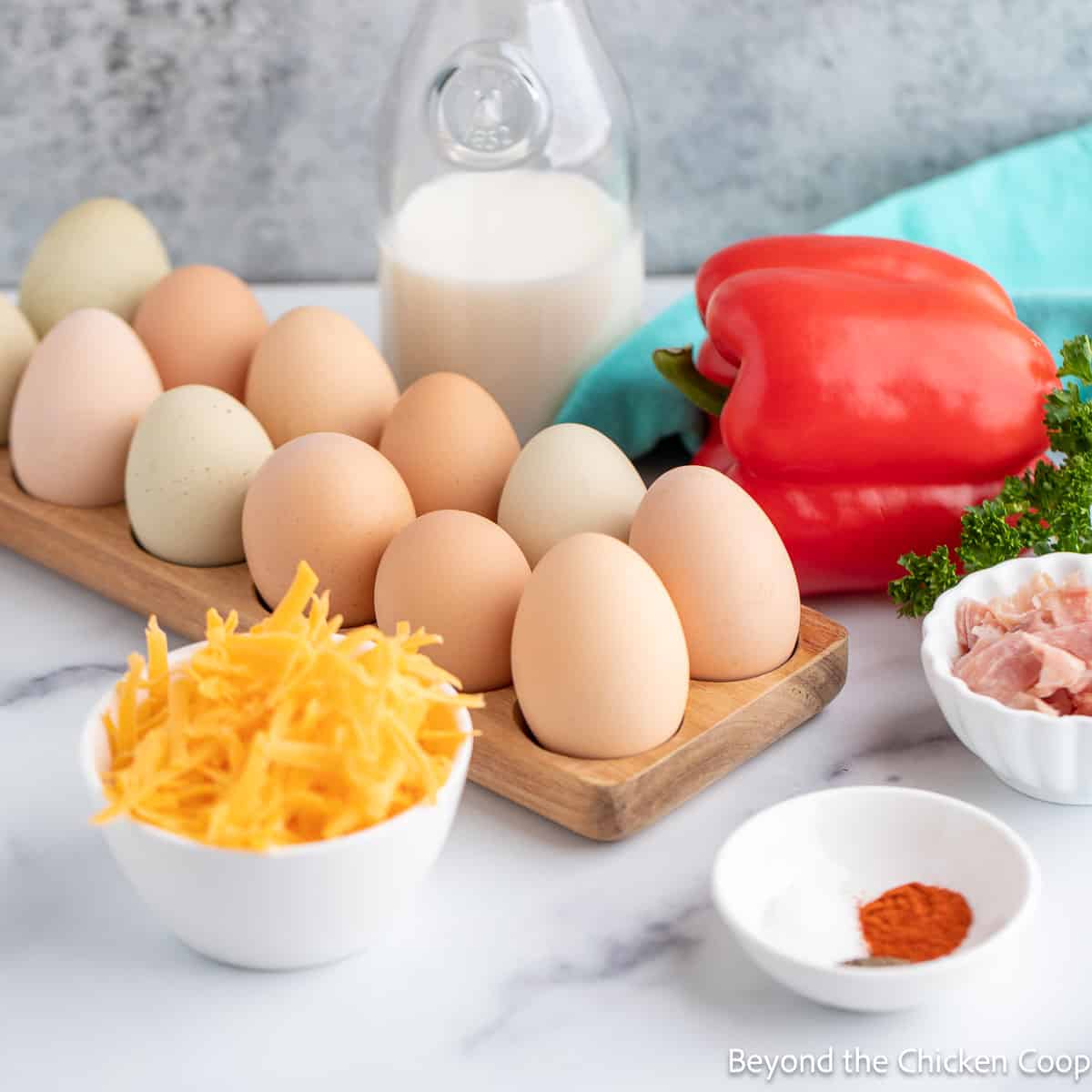 Fresh eggs next to a bowl of cheese, a bell pepper and chopped ham.