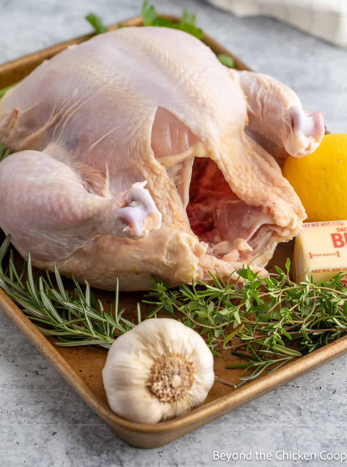 A whole raw chicken on a baking sheet with a lemon and fresh herbs. 