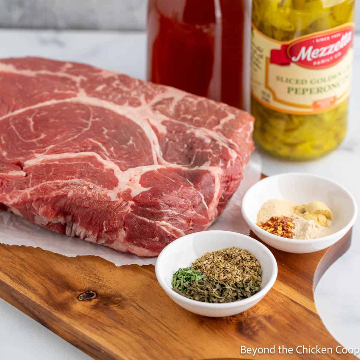 A chuck roast along with seasoning and pepperoncini peppers on a board. 