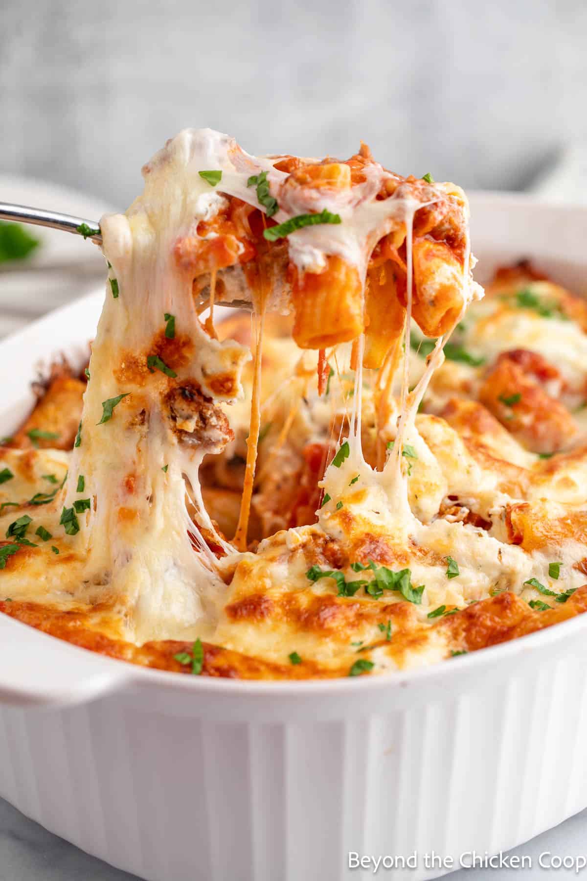 A large spoon lifting up a serving of pasta with the cheese stretching between the dish and the spoon. 