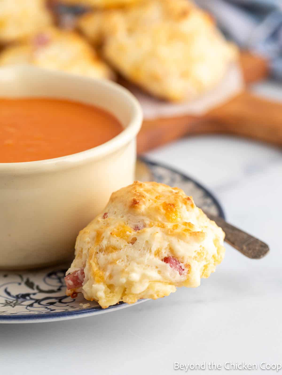 A biscuit with ham and cheese on a plate next to a bowl of soup.