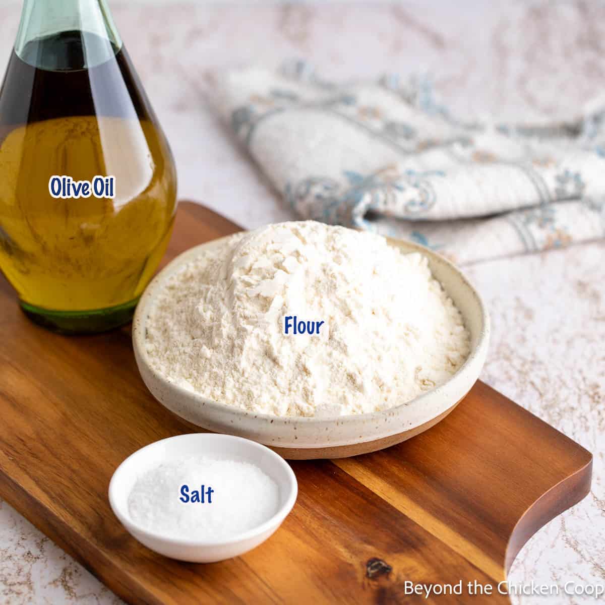 A bowl of flour, a small bowl of salt and a bottle of olive oil on a wooden board. 