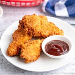 Three crispy chicken strips on a plate with a small bowl of barbecue sauce.