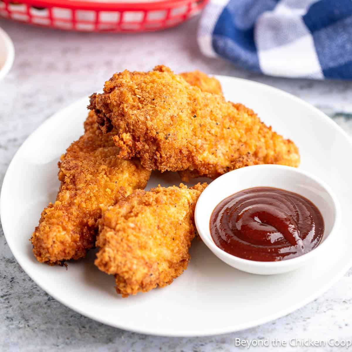 Three crispy chicken strips on a plate with a small bowl of barbecue sauce. 