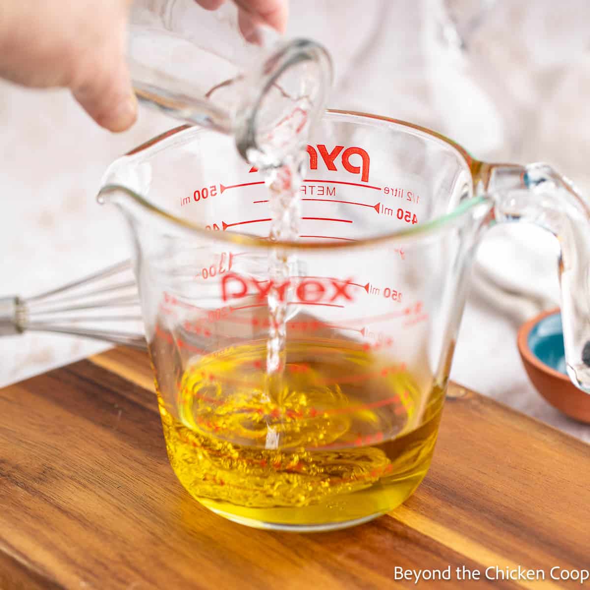 Pouring white wine vinegar into a glass measuring cup with olive oil in the cup. 