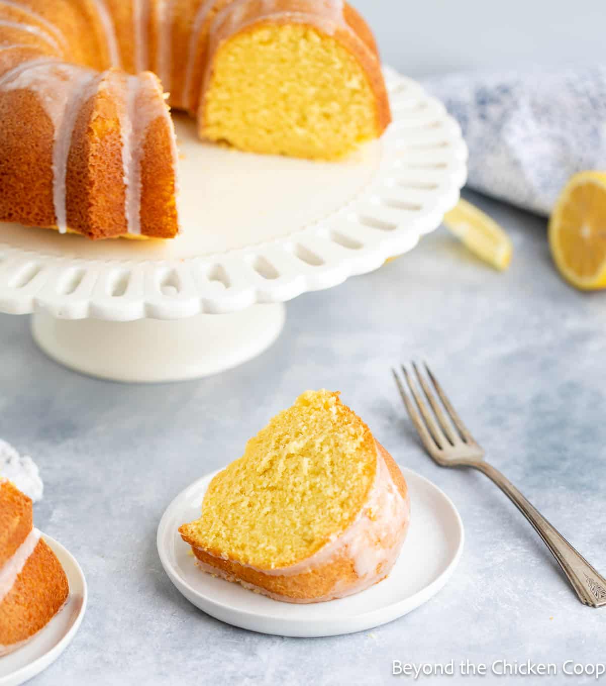 A lemon cake slice on a plate with the lemon cake on a cake platter. 