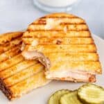 A grilled turkey sandwich on a plate next to sliced pickles.