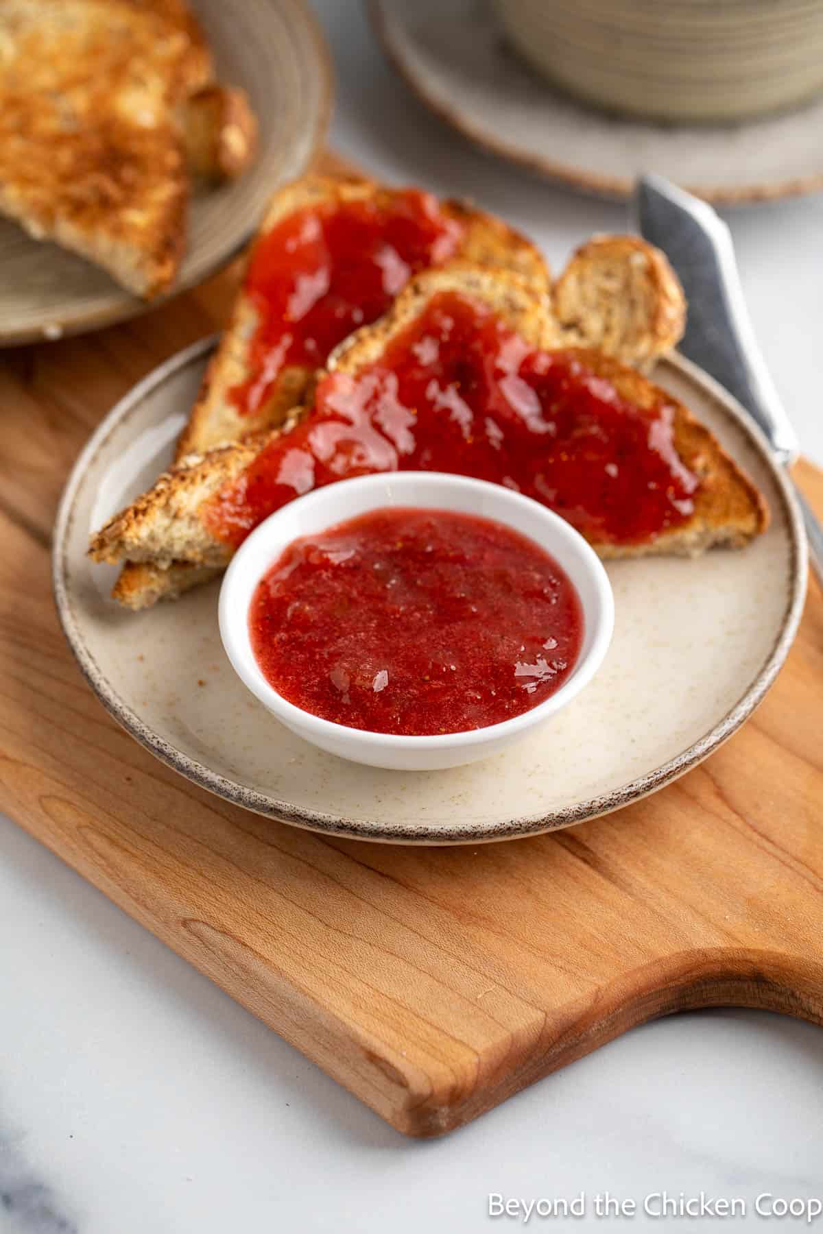 Homemade strawberry jam with rhubarb in a small bowl next to toast covered with the jam. 