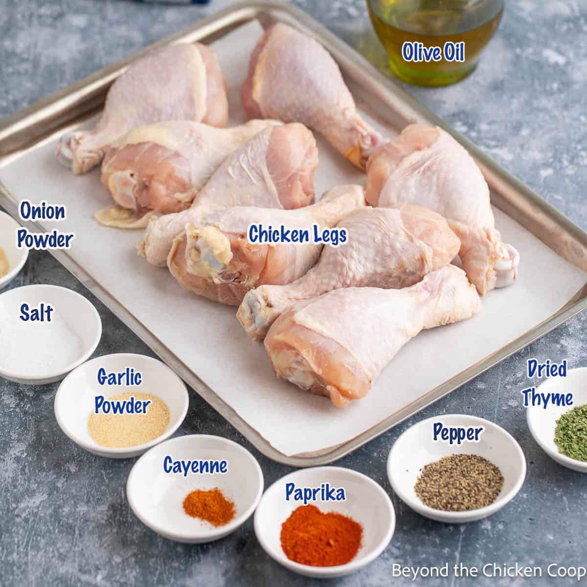 Raw chicken legs on a baking sheet next to small bowls filled with herbs and spices. 