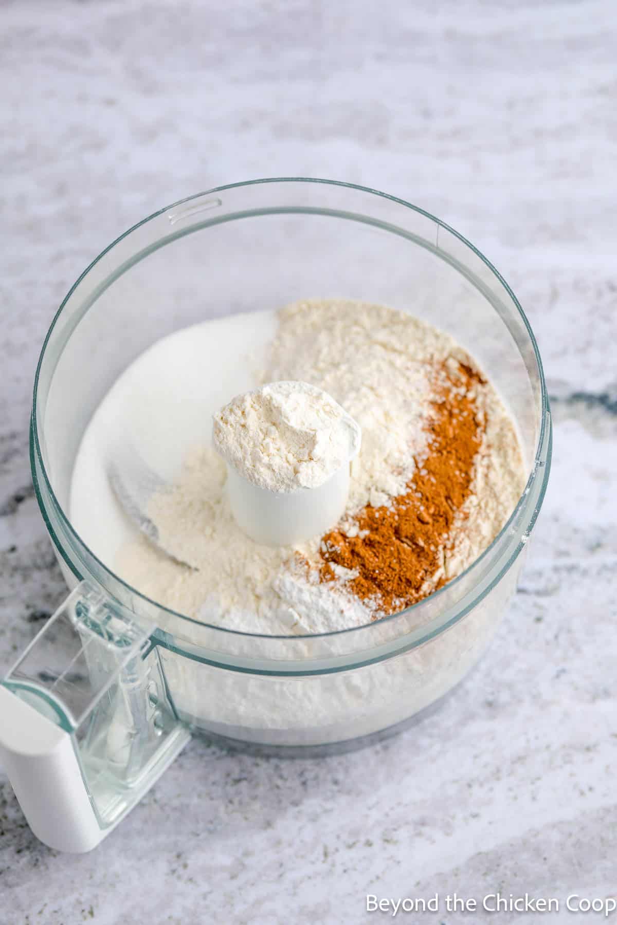 Flour, sugar, baking powder, and spices in a food processor bowl. 