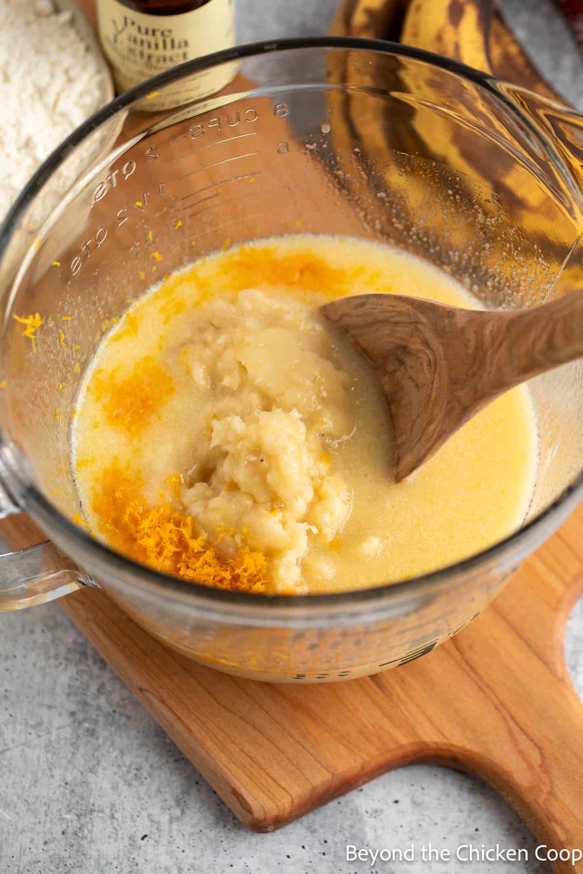 Mashed bananas and orange peel being added to a liquid mixture in a glass bowl. 