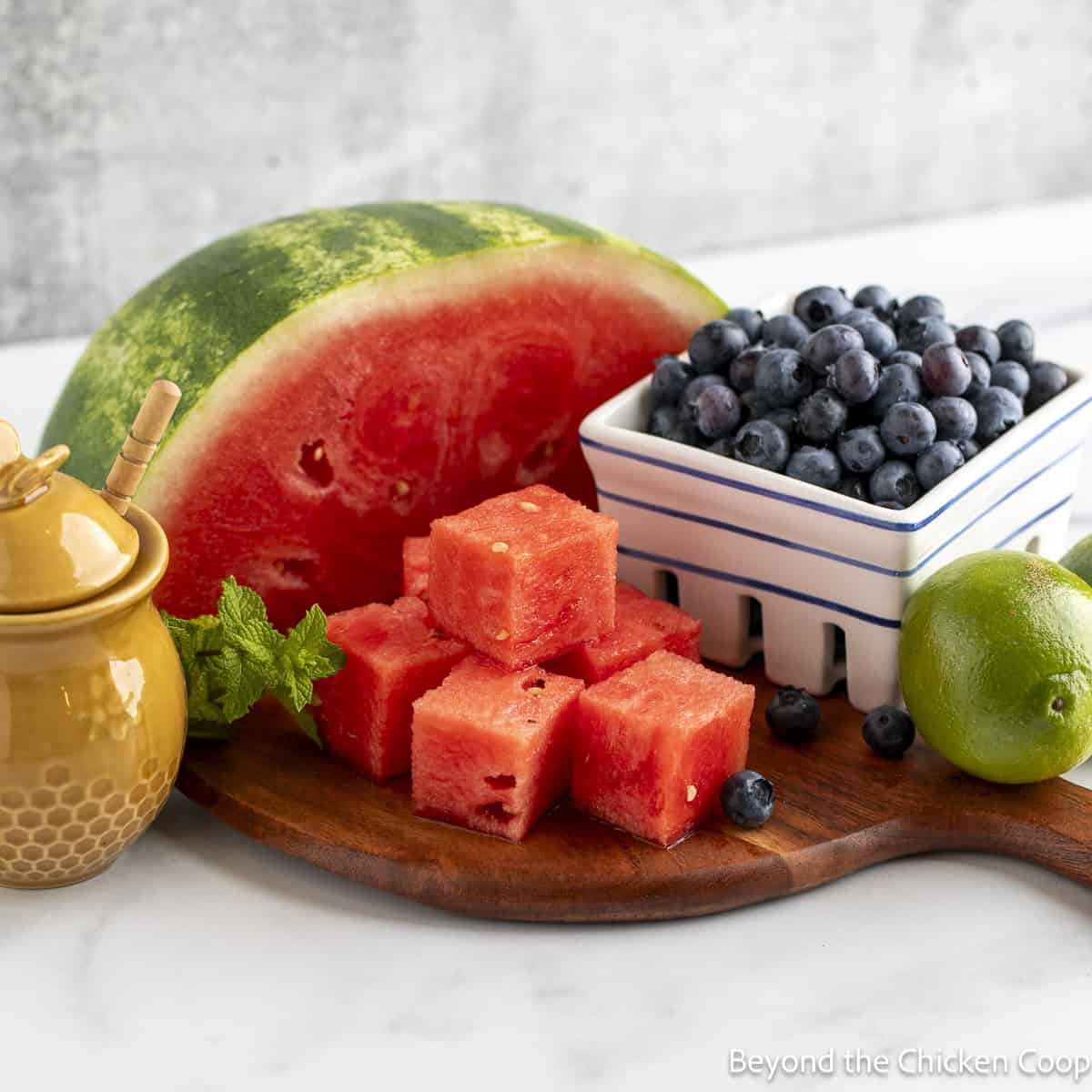 Fresh watermelon, a container of blueberries and a honey pot on a wooden board.