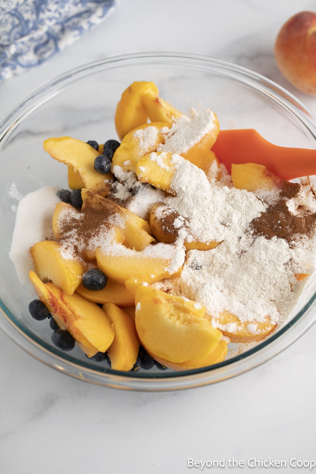Mixing sliced peaches, blueberries, flour, and spices together in a large glass bowl. 
