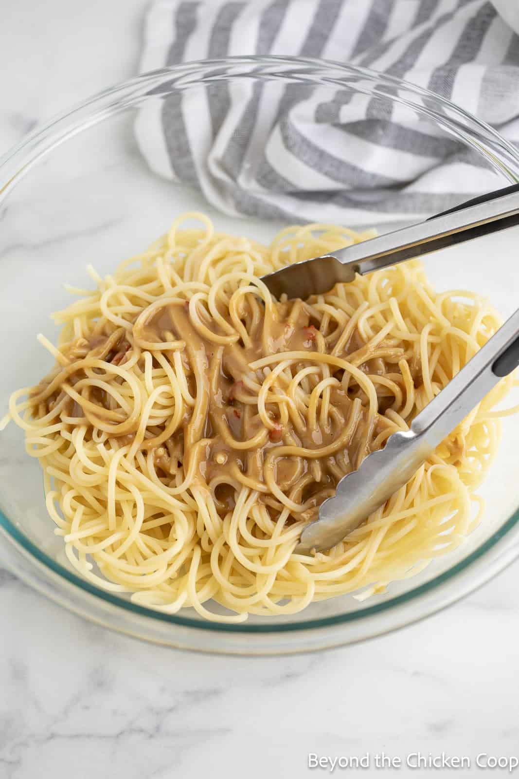Cooked spaghetti noodles being tossed with a peanut dressing. 