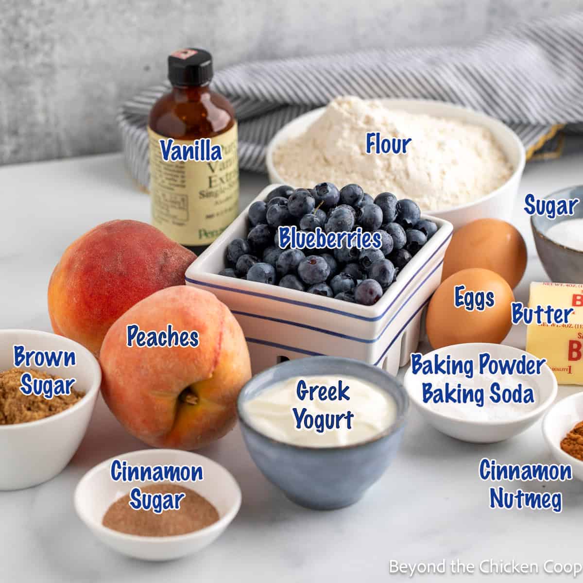 Blueberries, peaches, flour, eggs, butter, and other ingredients in small bowls.