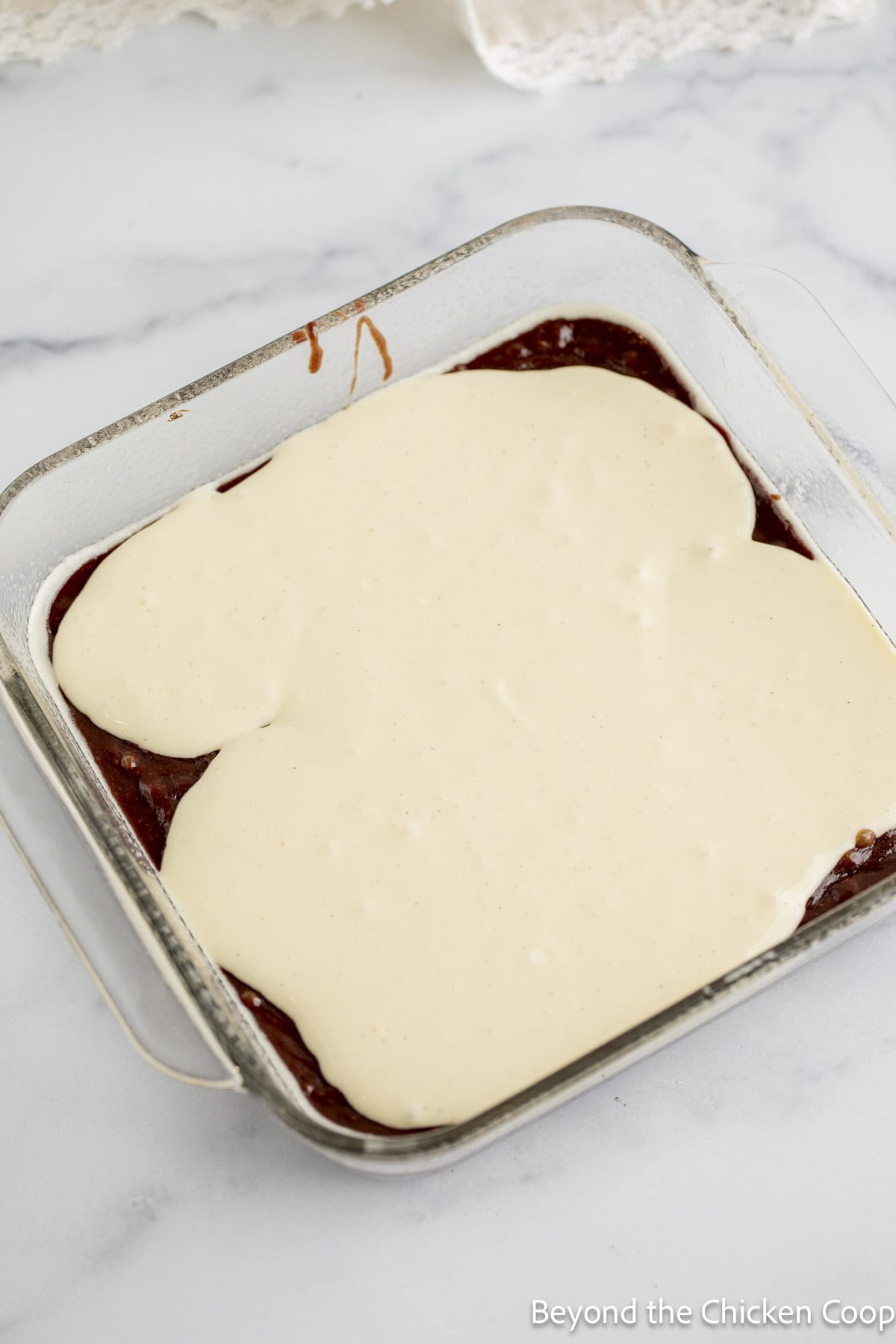 Cheesecake filling poured over brownie batter in a glass casserole dish.