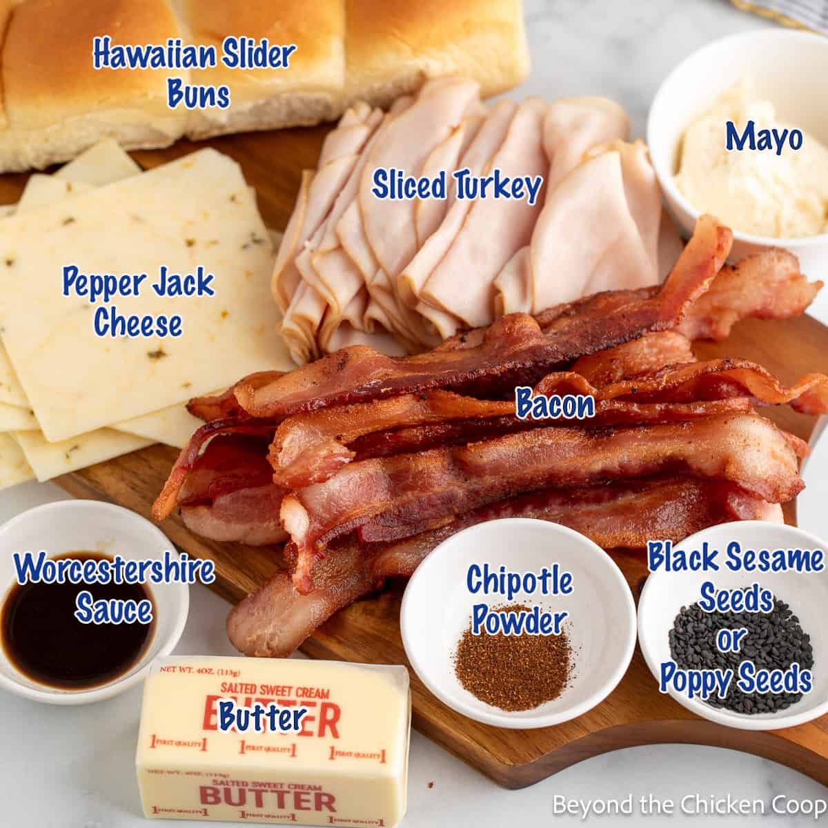 Sliced turkey, cheese, bacon, buns, and condiments on a wooden board.