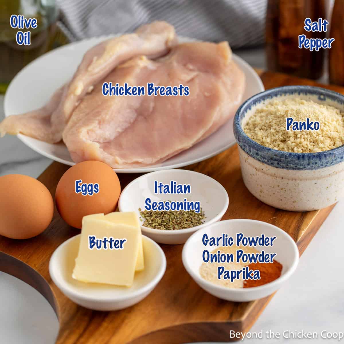 Chicken breasts, panko bread crumbs, eggs, butter, and seasonings arranged on a board.