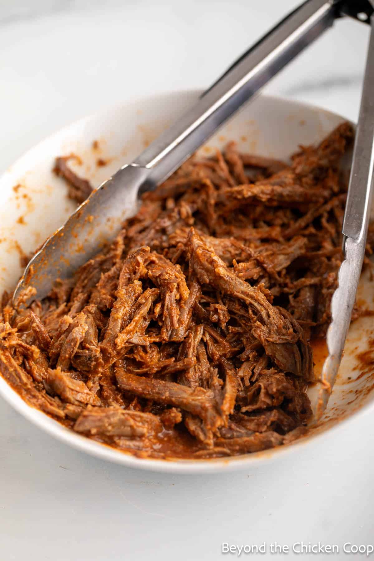 Shredded beef in a bowl with a pair of tongs.