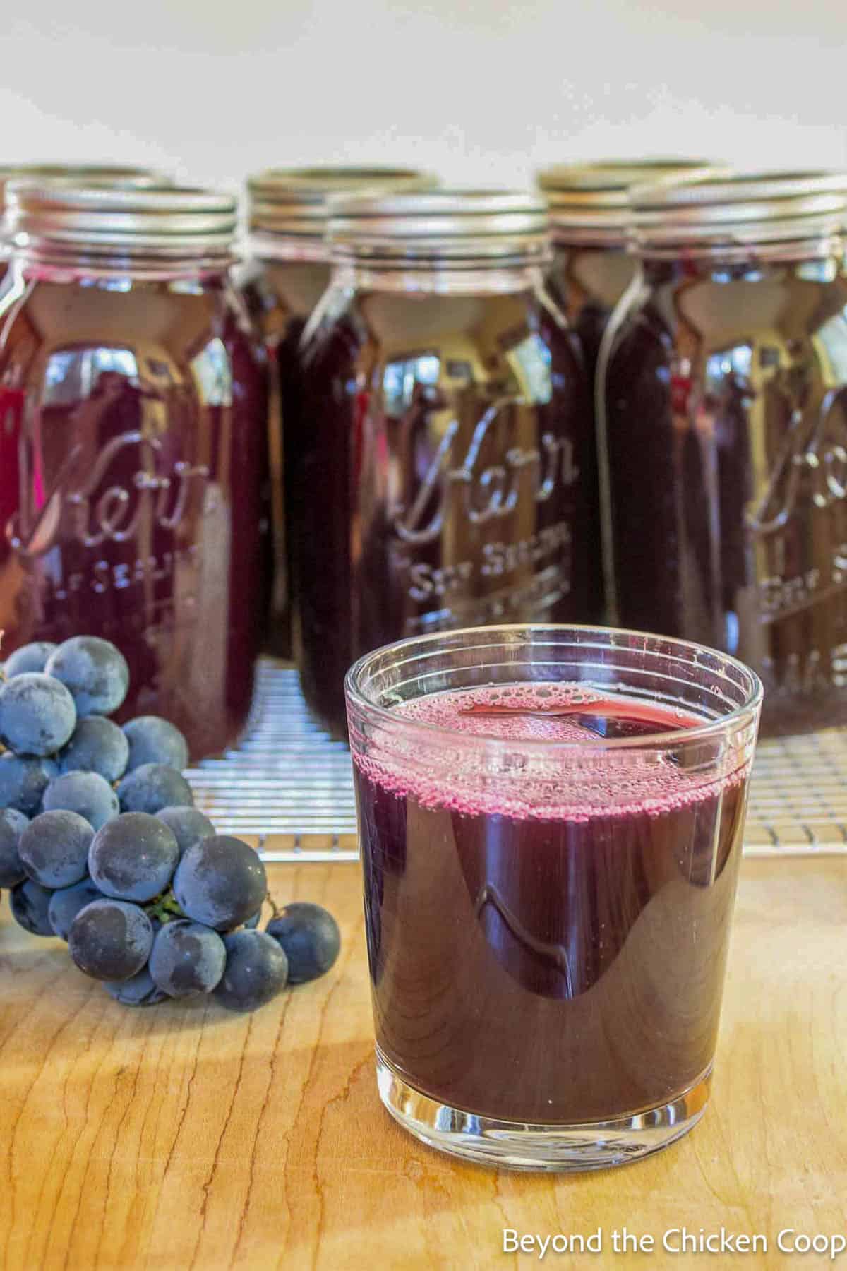 A glass of grape juice in front of jars of juice.