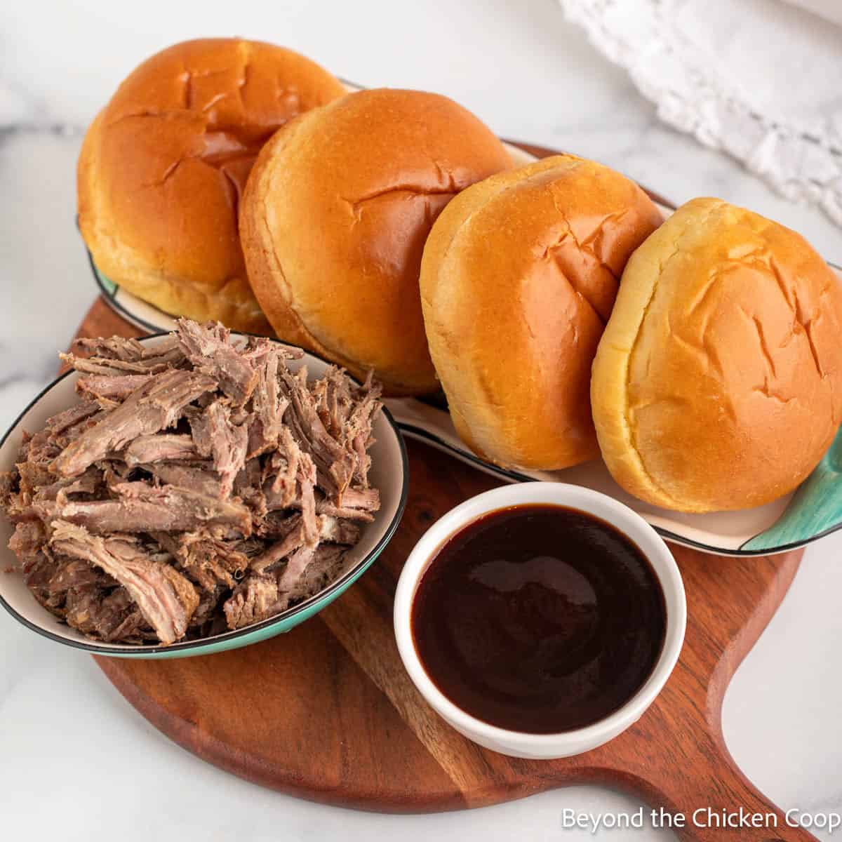 Shredded beef, a bowl of barbecue sauce, and hamburger buns on a round board.
